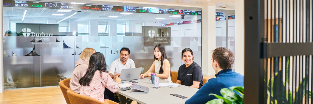 Students smiling around a table in Durham University Business School in front of a Bloomberg terminal