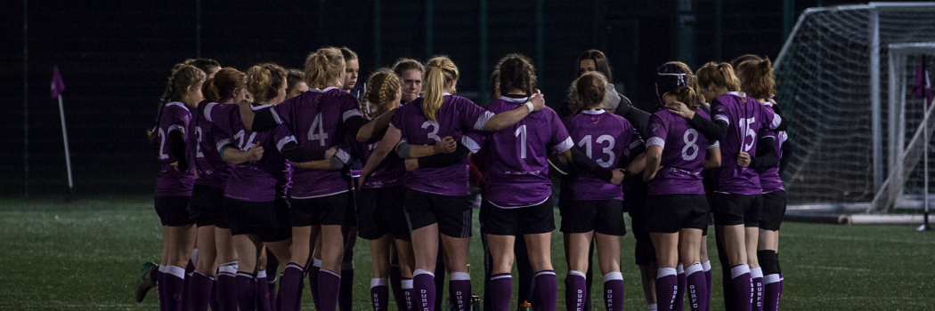 A huddle of Durham women's rugby players gather on the field ahead of a game.