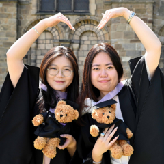 Two gowned graduates holding teddy bears and creating a heart shape with their arms