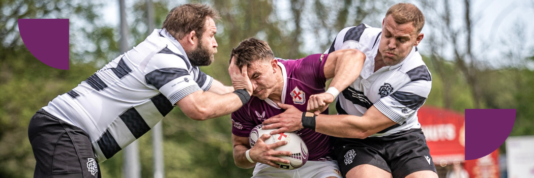 A group of three men playing rugby. The two men on either side of the man in the middle are on the same team and are tackling, trying to get the ball.