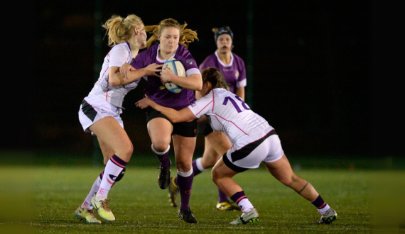 A female rugby player in purple runs through the middle of two other female players in white trying to tackle her.