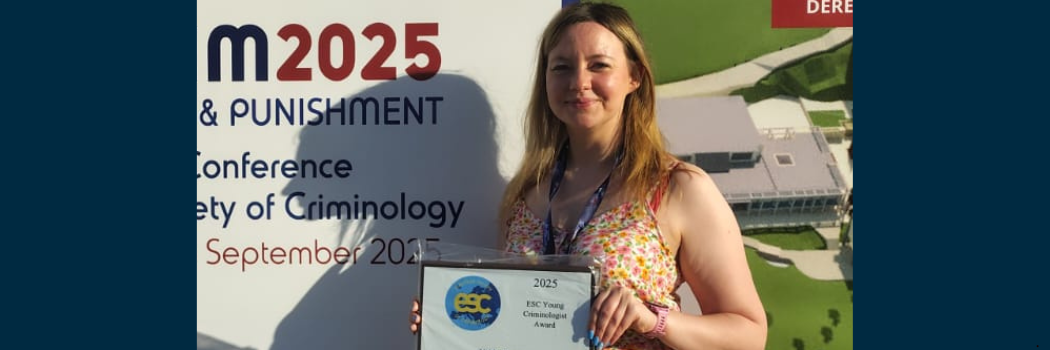 Dr Nikki Rutter faces the camera, in the sunshine, holding a framed award certificate with a conference banner in the background