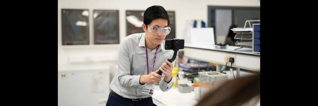 A student wearing safety goggles holds a phone, filming something off-shot