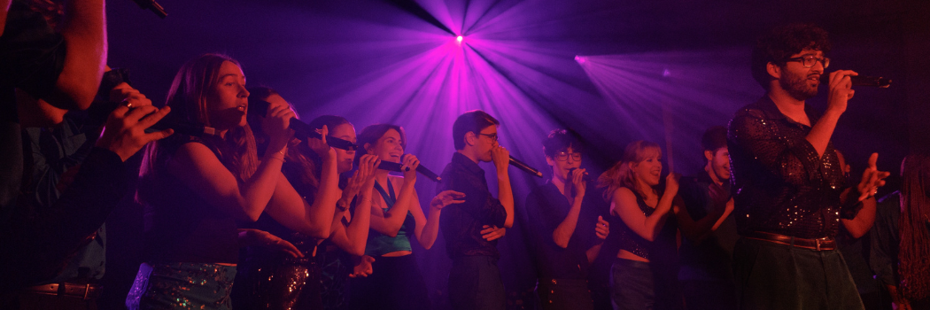A mixed semi-circle group of students singing into microphones lit up by purple lights.