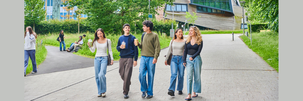 A group of students walk along a path with trees and wood panelled building behind them.