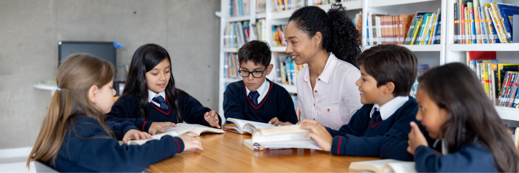 A group of children in school uniform sitting round a table reading with a female teacher in a white shirt