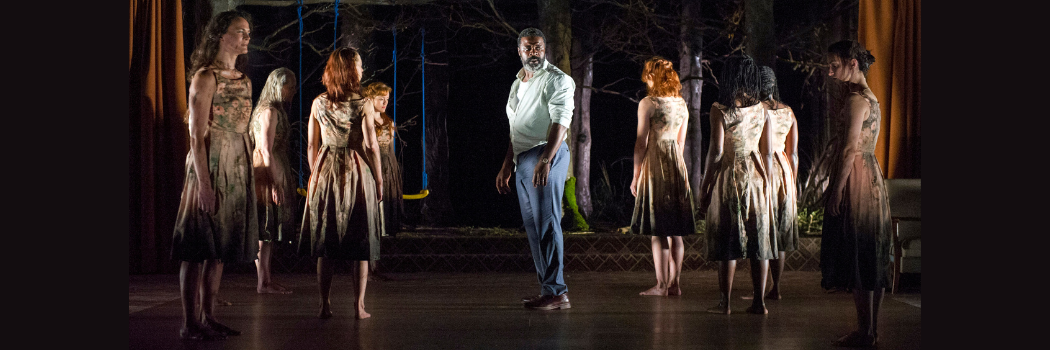 Actors on a shadowy stage, with a man in the middle and women wearing dresses stood either side of him
