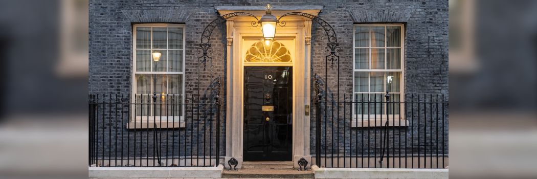 A photograph of the black door to Number 10 Downing Street. A light shines from the centre of the black wrought iron archway in front of the door.