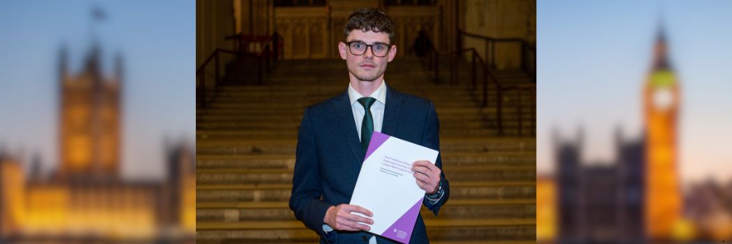 A man wearing a suit, shirt and tie, and wearing glasses, looks at the camera while holding a report.