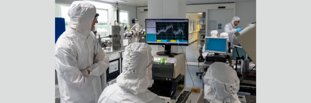 Three scientists in white overalls and hoods look at a graph on a computer screen.