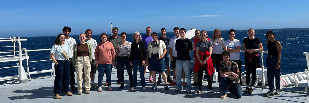 A group of men and women dressed casually smiling on the deck of a ship with bright blue sky and dark blue ocean in the background.