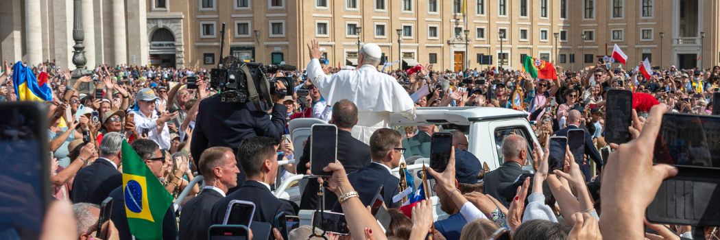 As seen from behind, Pope Leo waves to crowds while standing on the back of the Popemobile.