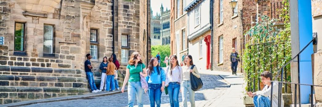 A group of female students walk down a cobbled street in the sunshine. Other students are either side of the group. In the background are the towers of Durham Cathedral.
