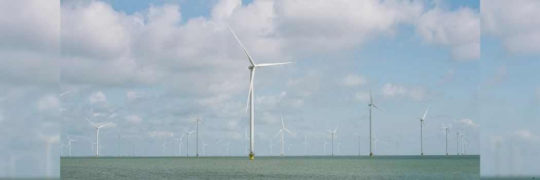An offshore wind farm stands in a grey sea with a blue and cloudy sky behind.
