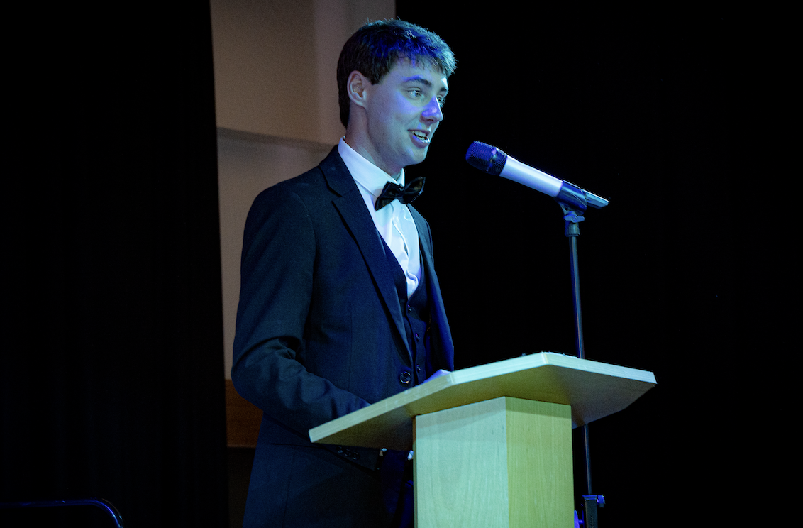 A young man, our 23/24 JCR Chair, Max Bamford, in a black tuxedo speaks at a podium during the John Snow College awards ceremony. He stands under stage lighting, addressing the audience with a microphone against a dark curtain backdrop.