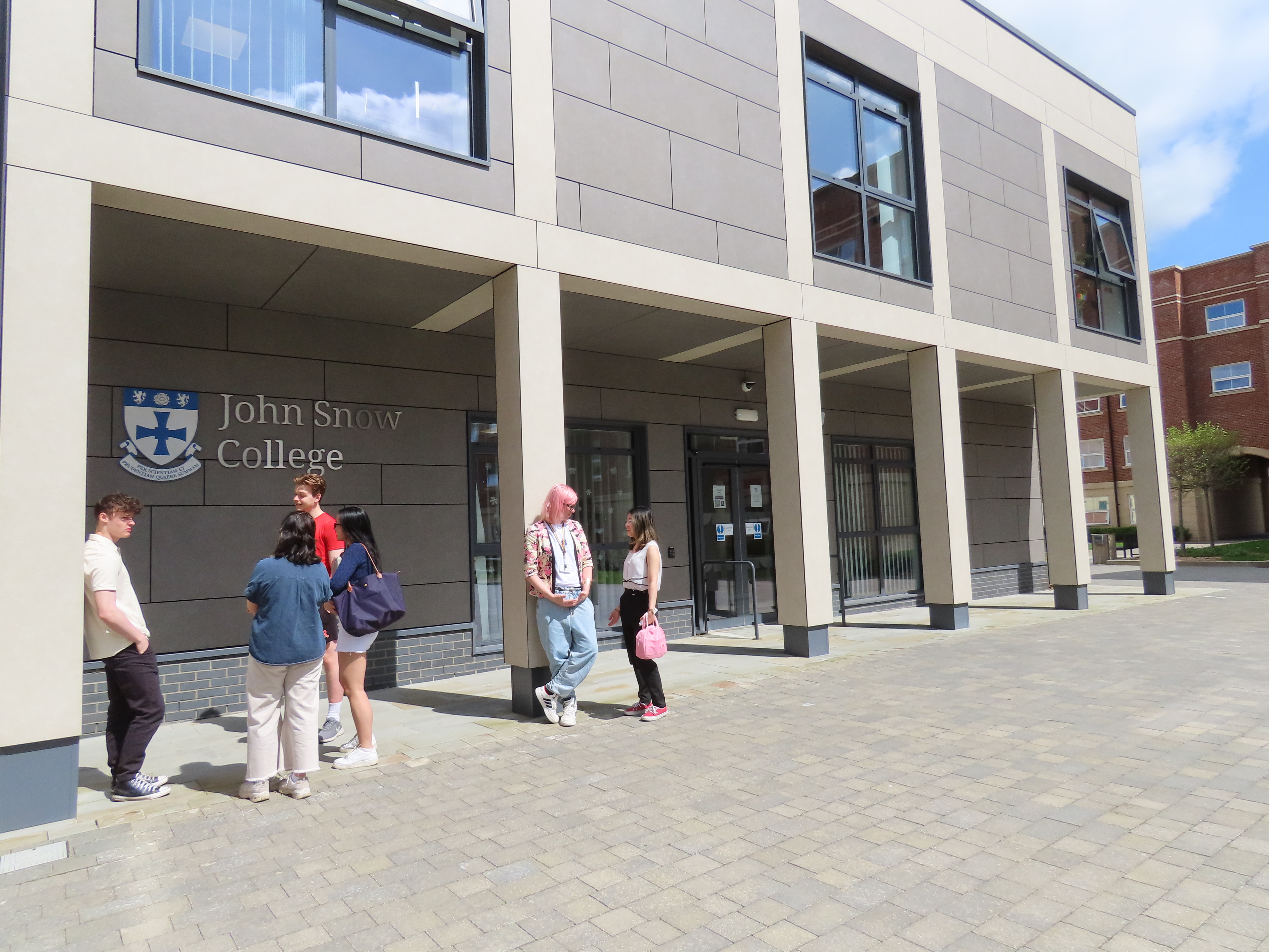 A group of students stand and chat outside the entrance to John Snow College on a sunny day. The modern building features large windows, a light stone façade, and the college crest mounted on the wall.