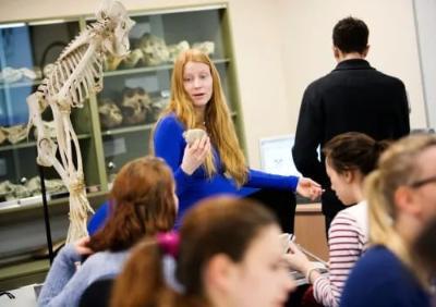 Fire Kovarovich sits next to a teaching skeleton of a primate, speaking to students gathered in a laboratory. In the background, there are glass-fronted cabinets with casts and fossils of  primate skulls.