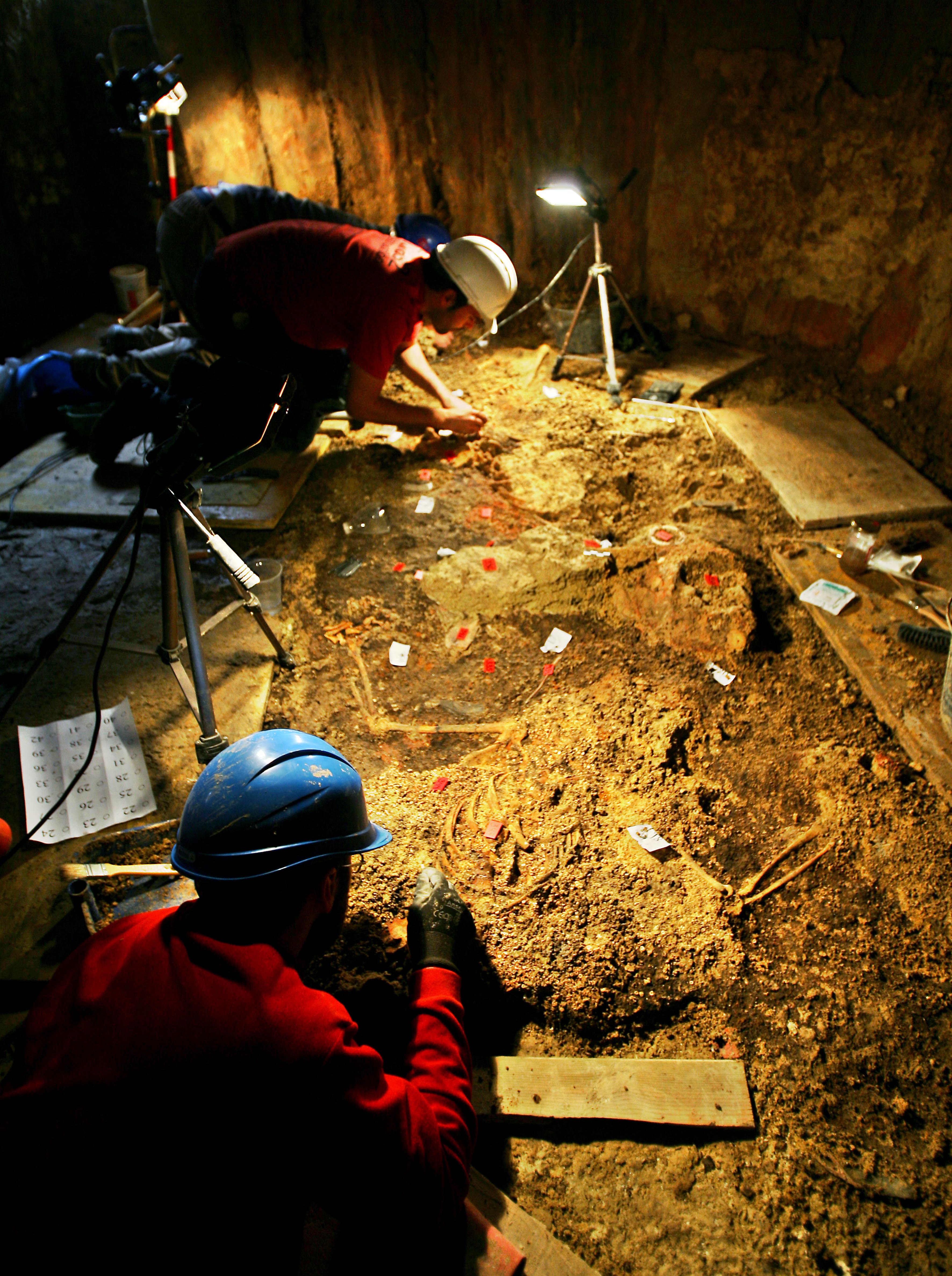 Three archaeologists in protective gear excavating an ancient burial site, with aid from large lamps.