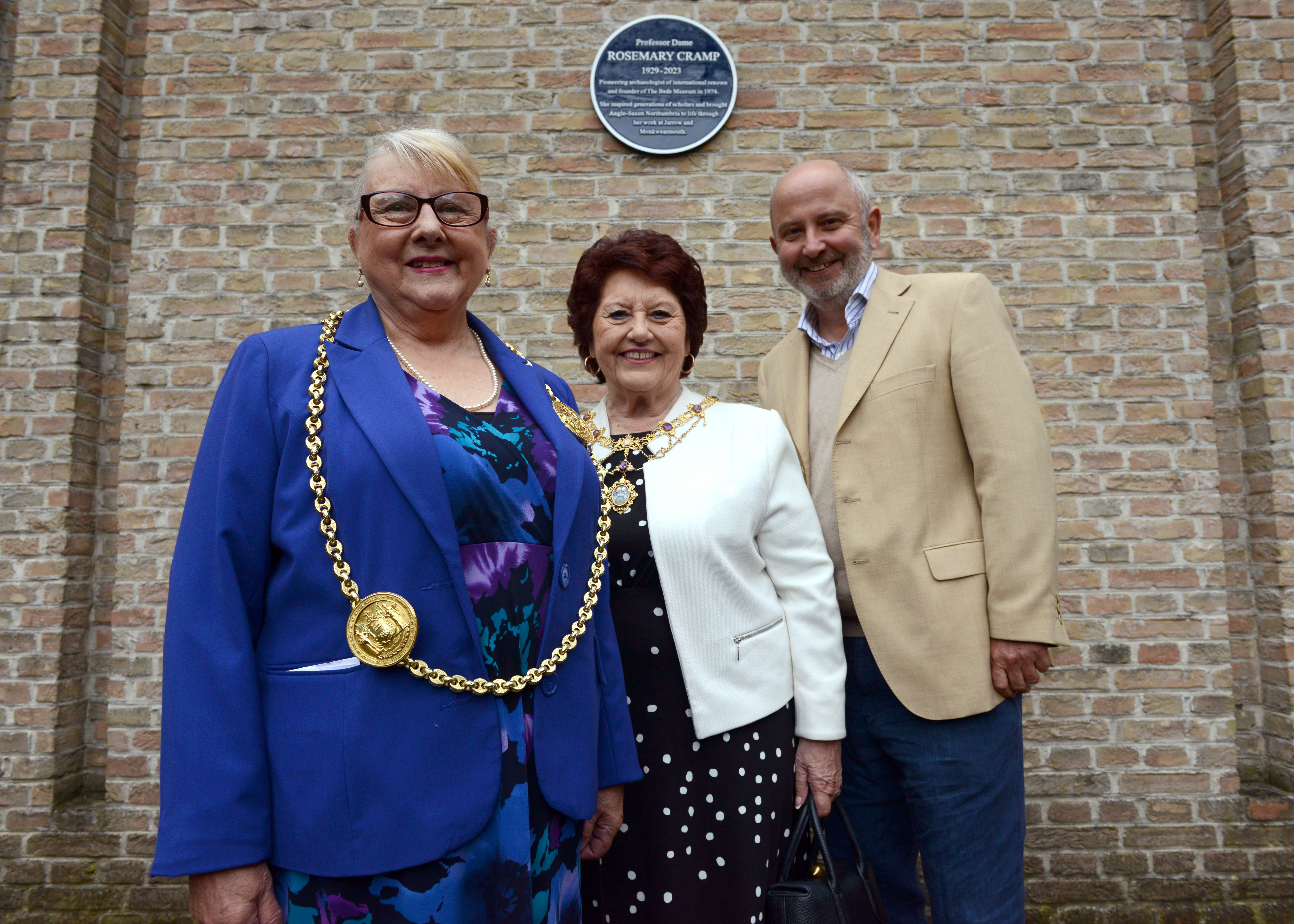 Three smartly dressed and smiling people, Councillor Fay Cunningham, Mayoress Stella Matthewson and Professor Chris Gerrard, stand in front of a wall that features a blue plaque honouring Professor Dame Rosemary Cramp, 1929 - 2023.