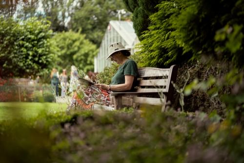 A woman in a hat is sitting on a bench in a garde reading.