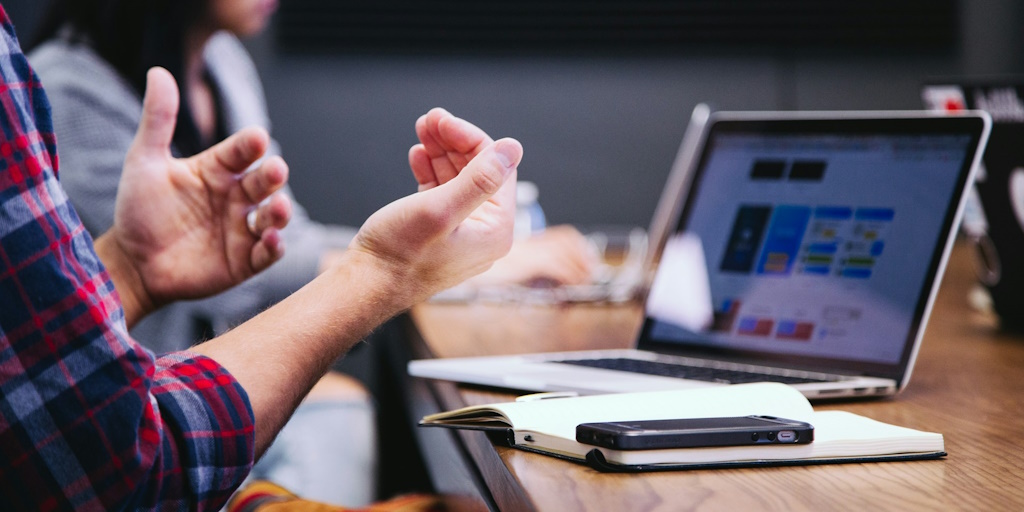 Closeup of a mans hands gesturing while talking with a laptop infront of him. People are sat around a meeting table.