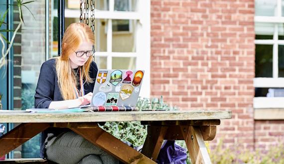 oung woman with long auburn hair studies her laptop as she sits outside a building at St John's College. It is a sunny day and she wear a long sleeved black top.