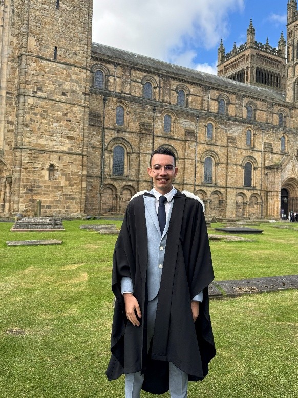 Georgios stood in graduation gown in front of Durham Cathedral