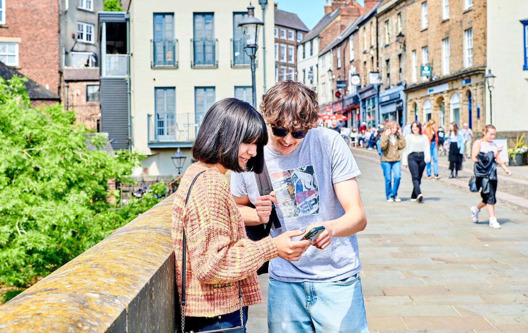 Two students looking at a phone in Durham City on a bridge