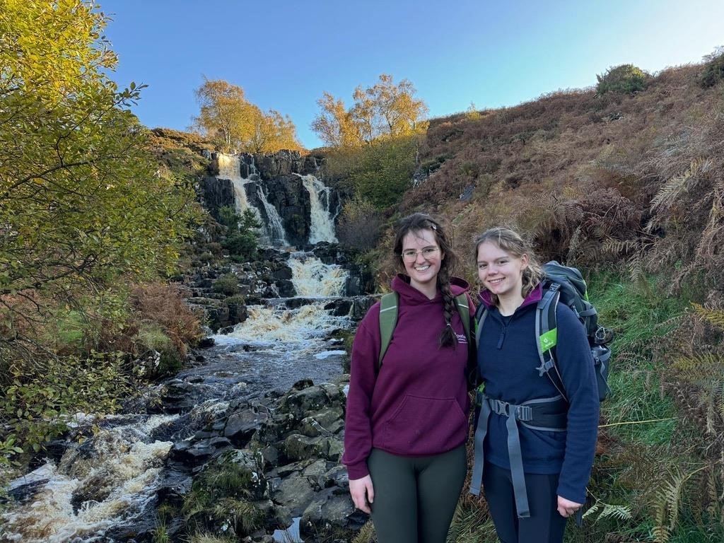 Two students in hiking gear pose in front of a small waterfall in the countryside.