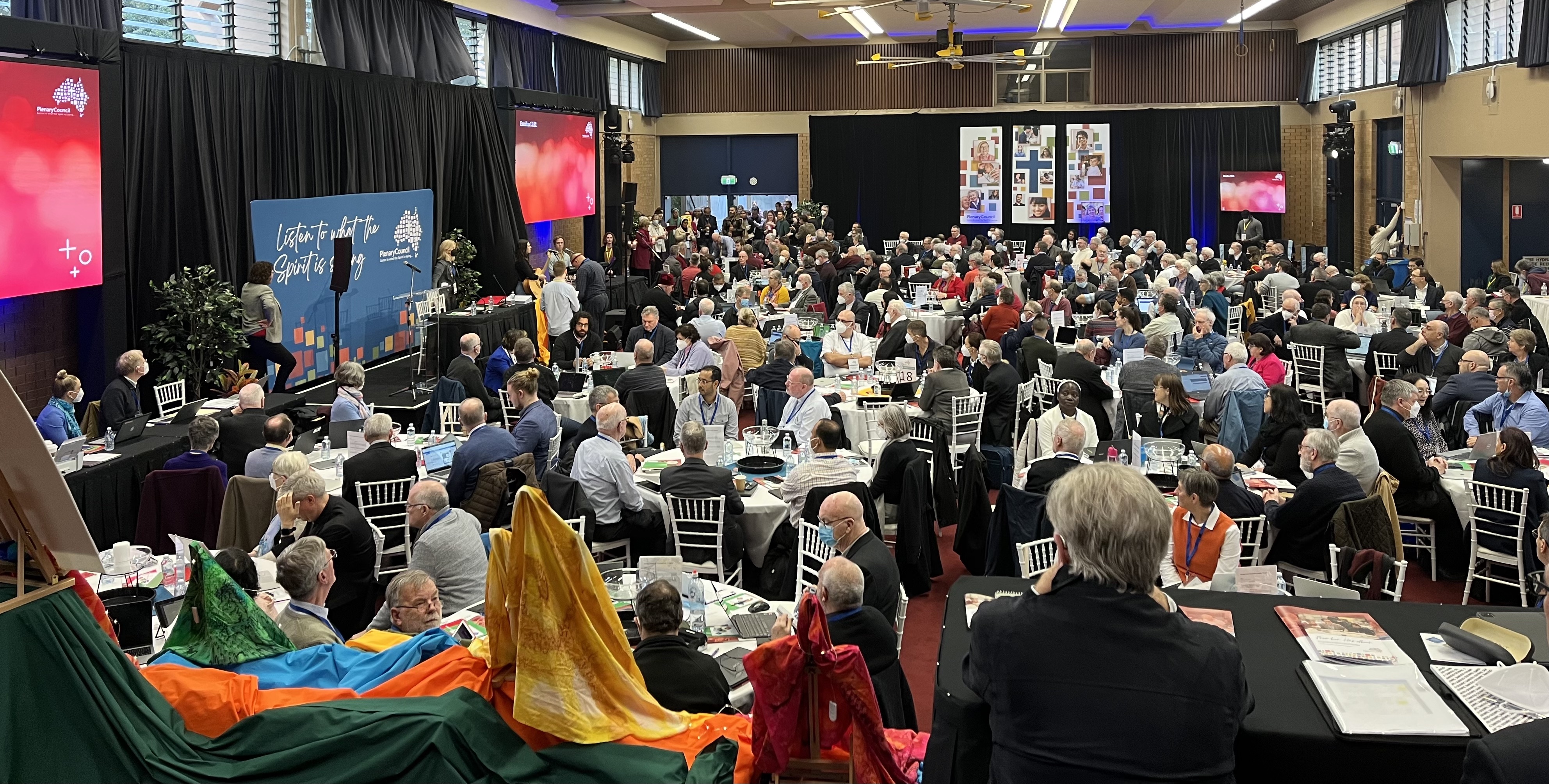 A room full of people sitting at round tables with a sign on stage saying