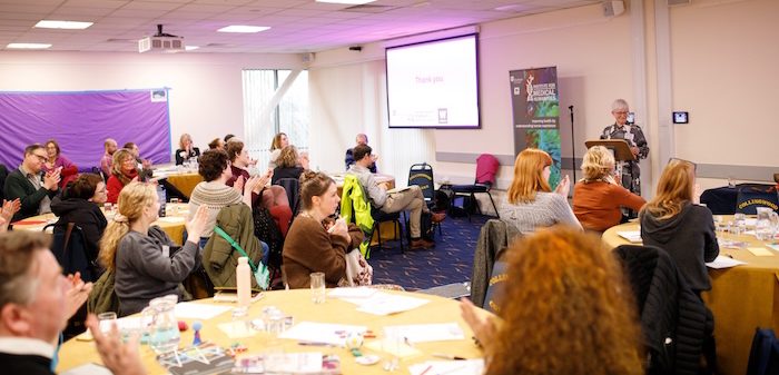 A conference room filled with attendees seated at round tables, listening to a speaker at a podium. The room is brightly lit and the audience appears engaged.