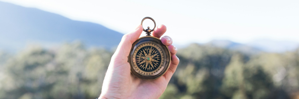 hand holding a brass compass against a backdrop of sky and mountains