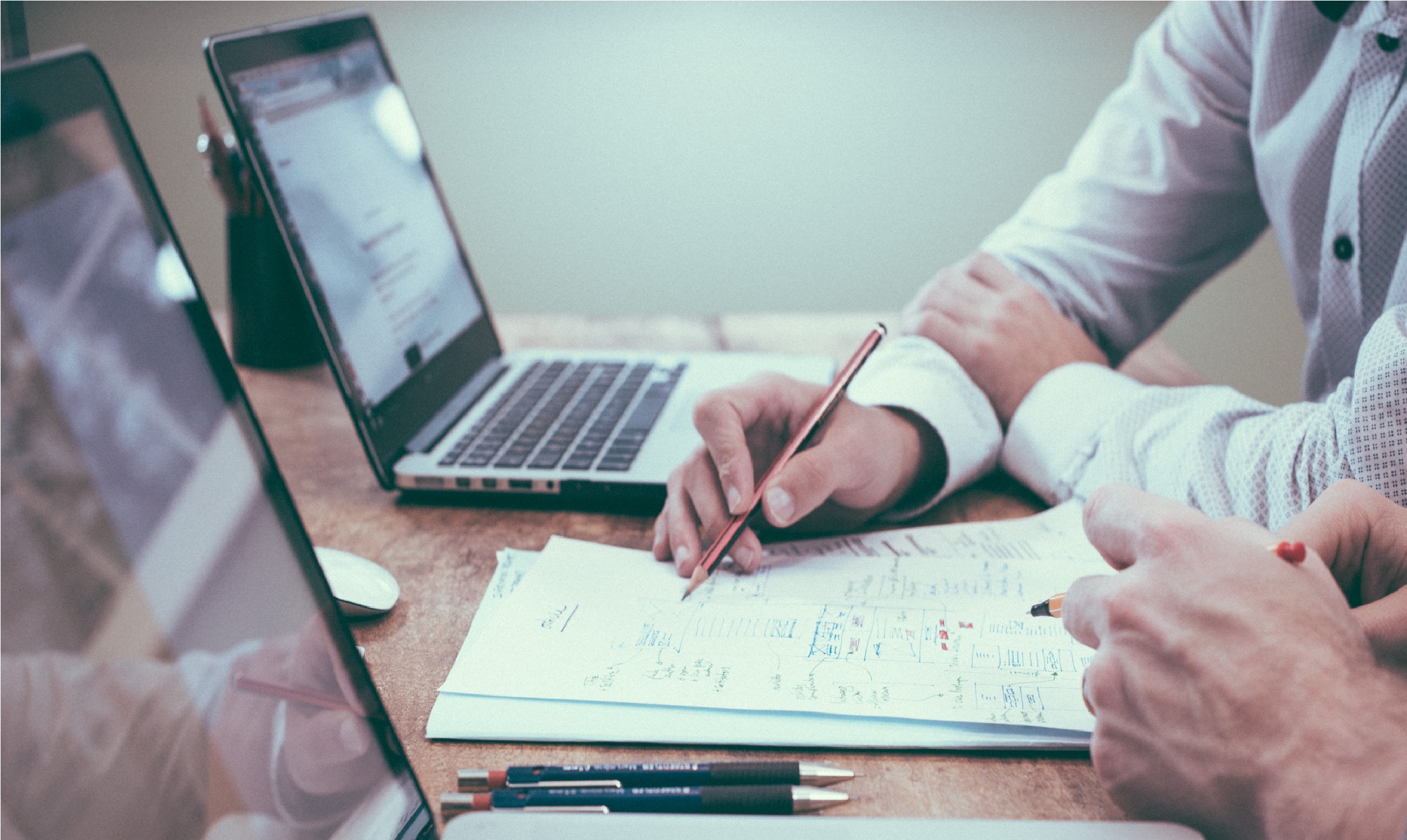 Two laptops open on a desk in front of two people's hands and some unintelligible paperwork.