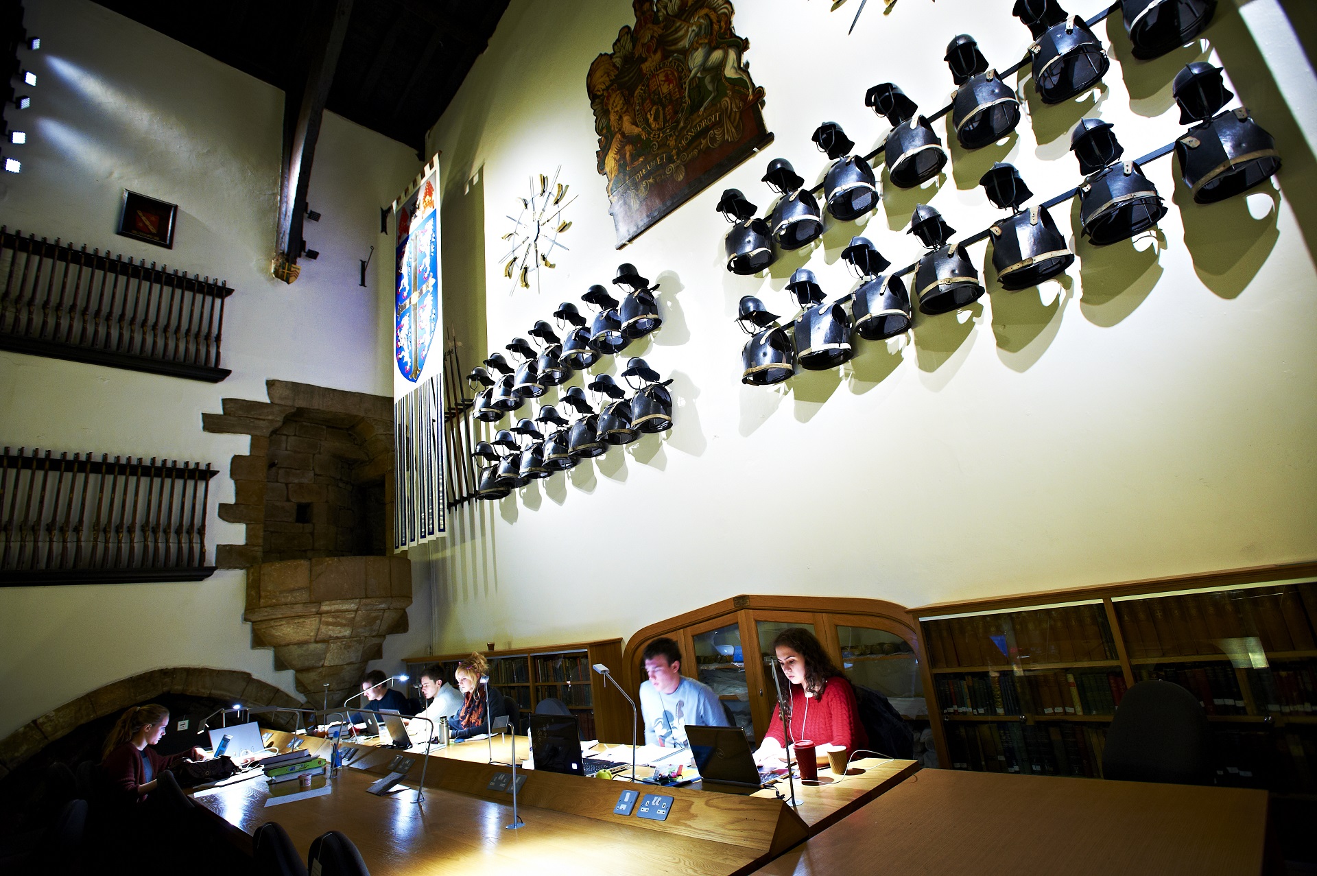 A coloured photograph taken in the Minstrel’s Gallery, looking towards the south wall of the Great Hall. At the top of the image are the armour. Below students are studying.
