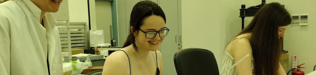 Two Work Experience participants sit at a table in a Conservation studio with historic documents and modern forms on the table. A member of the Conservation team stands next to them smiling.